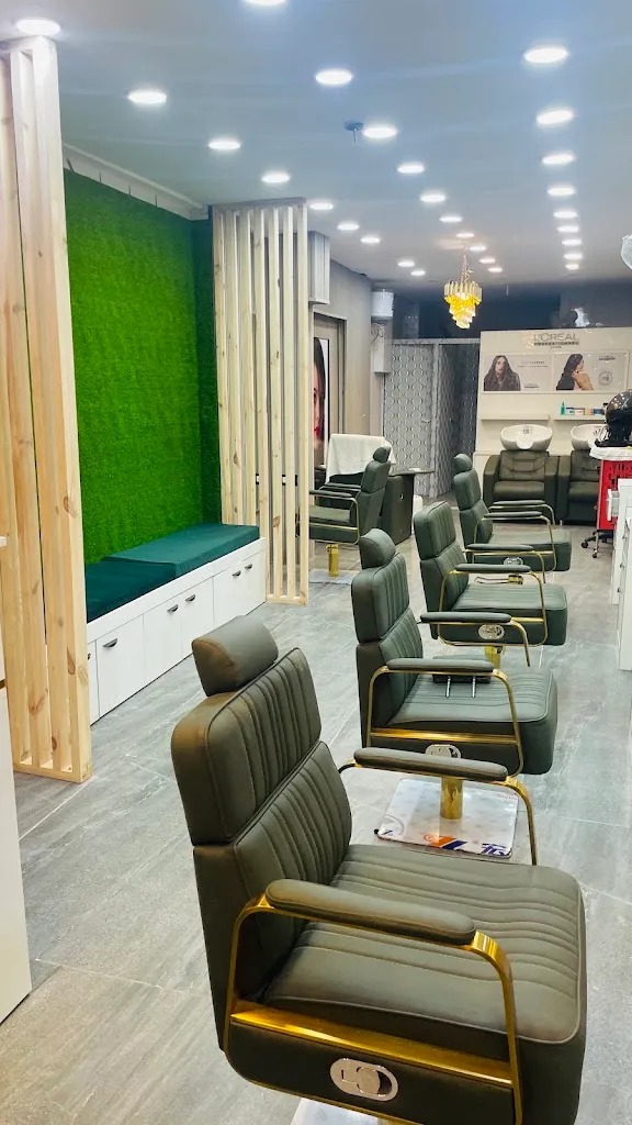 A row of styling chairs with gold-toned metal frames and dark upholstery, facing a mirrored wall panel. An artificial-grass accent wall and a shelf of L'Oreal Professionnel haircare products stand at the back of the room.